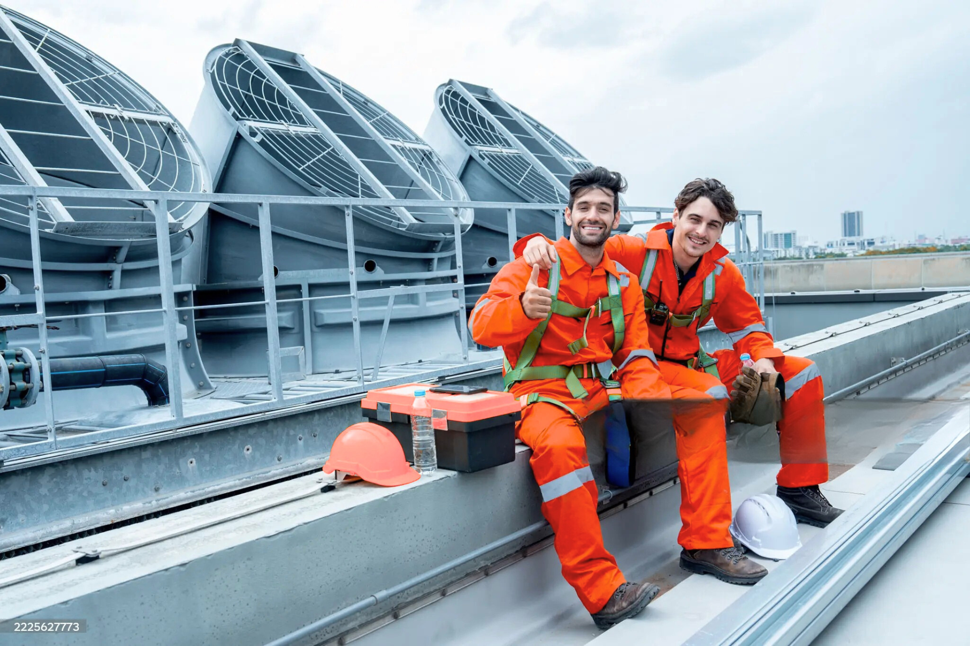 "Two workers in orange safety suits and helmets, sitting on a rooftop with large industrial fans in the background. One worker is giving a thumbs-up while the other is smiling. There are tools and a water bottle beside them."