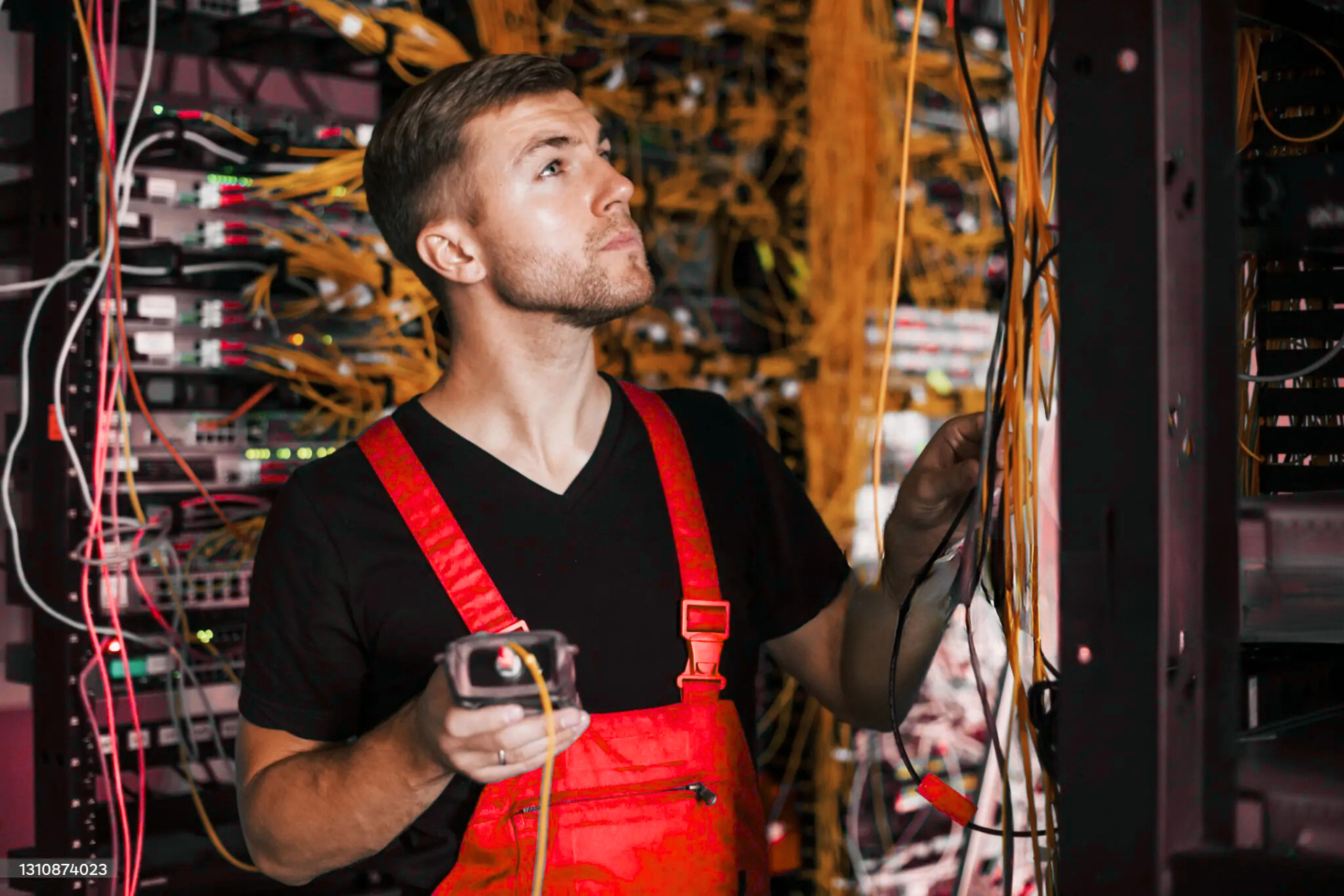 "An electrician in a red work bib standing in front of a server rack, holding a tool in one hand and inspecting cables with the other, surrounded by a tangle of yellow and black wires."