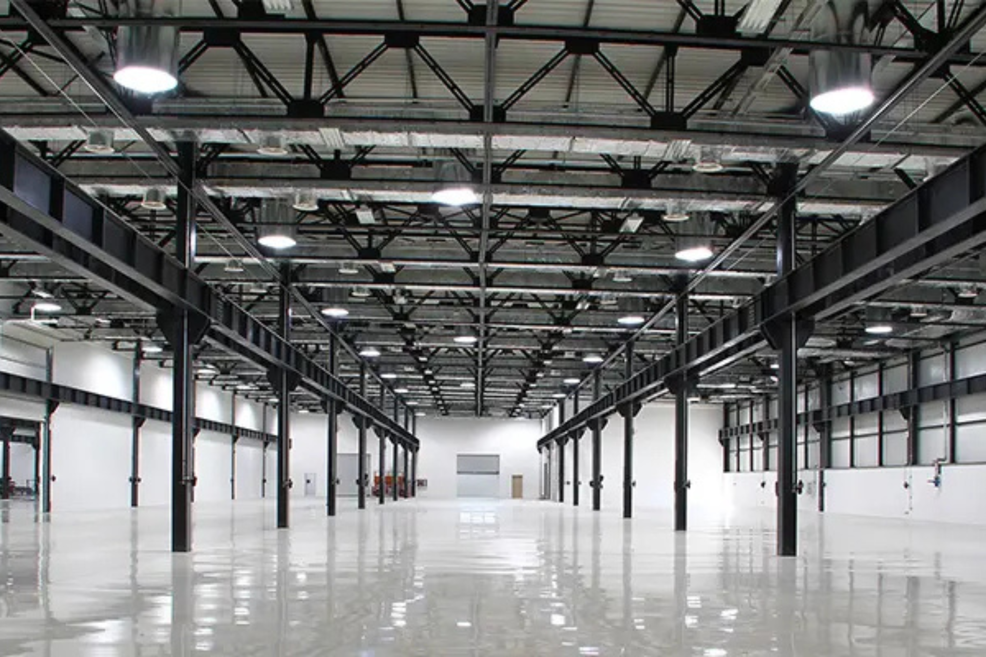 Interior of a large, empty industrial warehouse with polished white floors, high ceilings, exposed steel beams, and bright overhead lighting.
