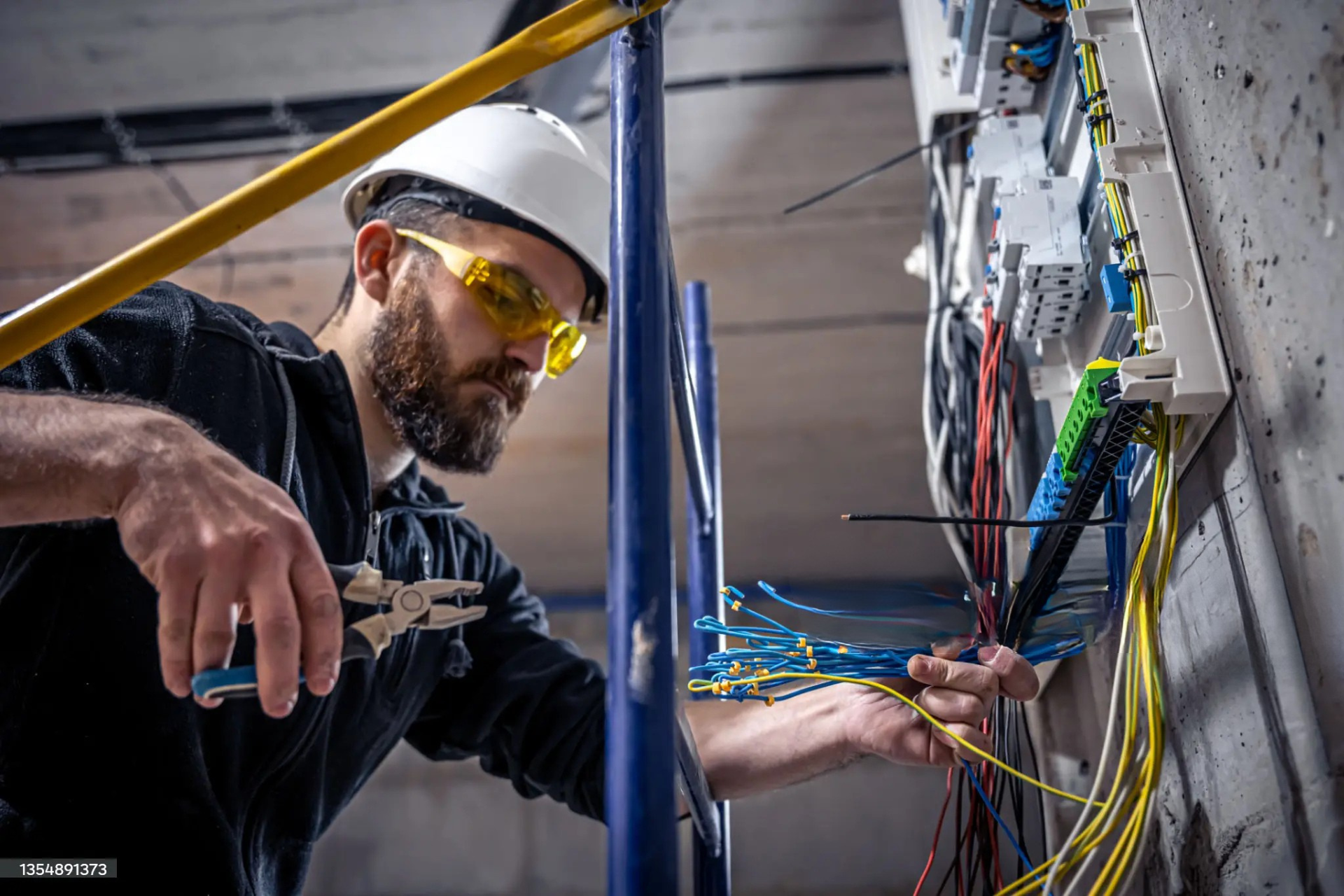 "An electrician wearing a helmet and safety glasses, working with electrical wires and using pliers to organize and connect wires in a circuit panel."