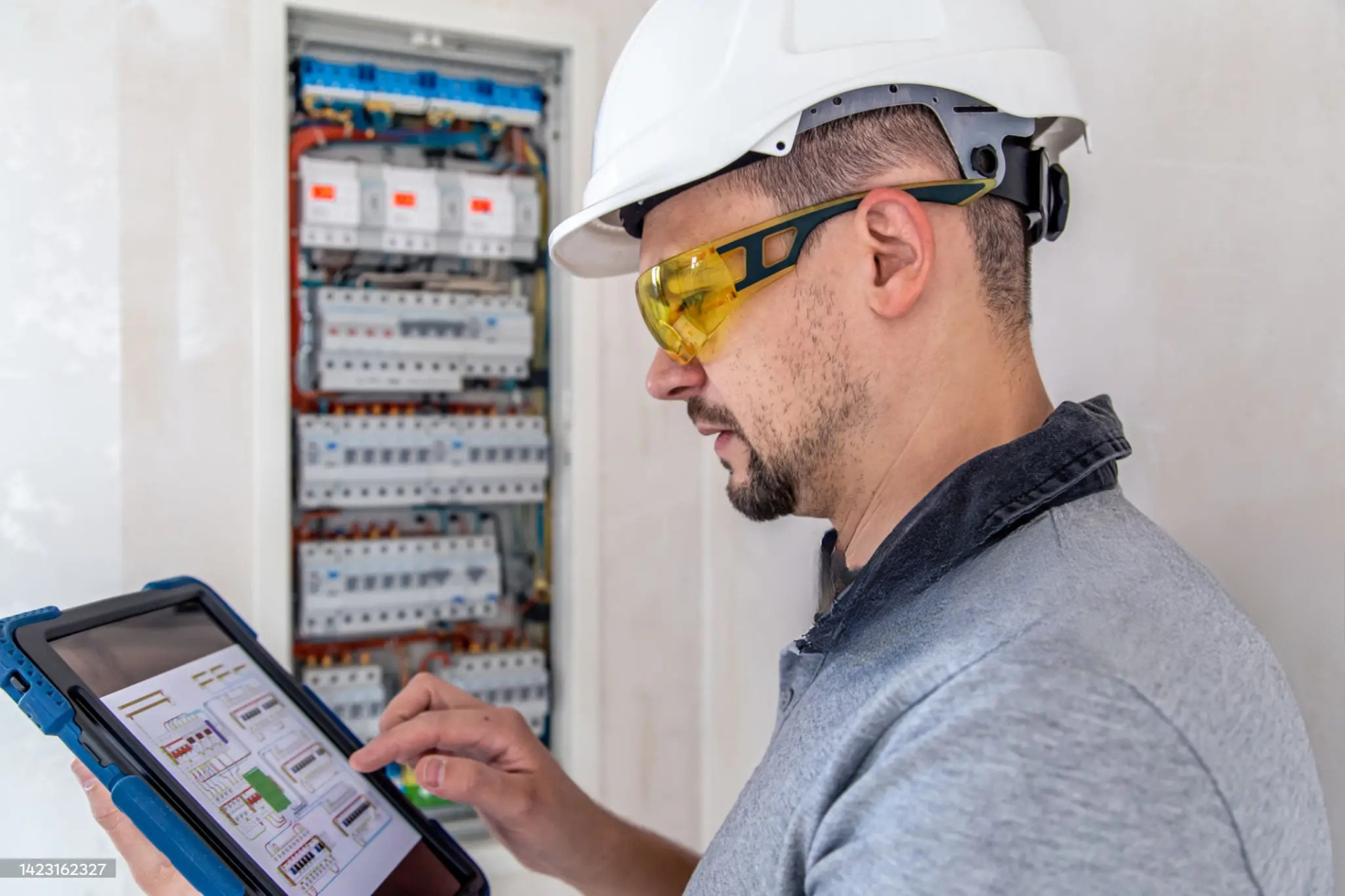 "Electrician wearing a white hard hat and yellow safety glasses, using a tablet to check electrical system diagrams next to a circuit breaker panel."