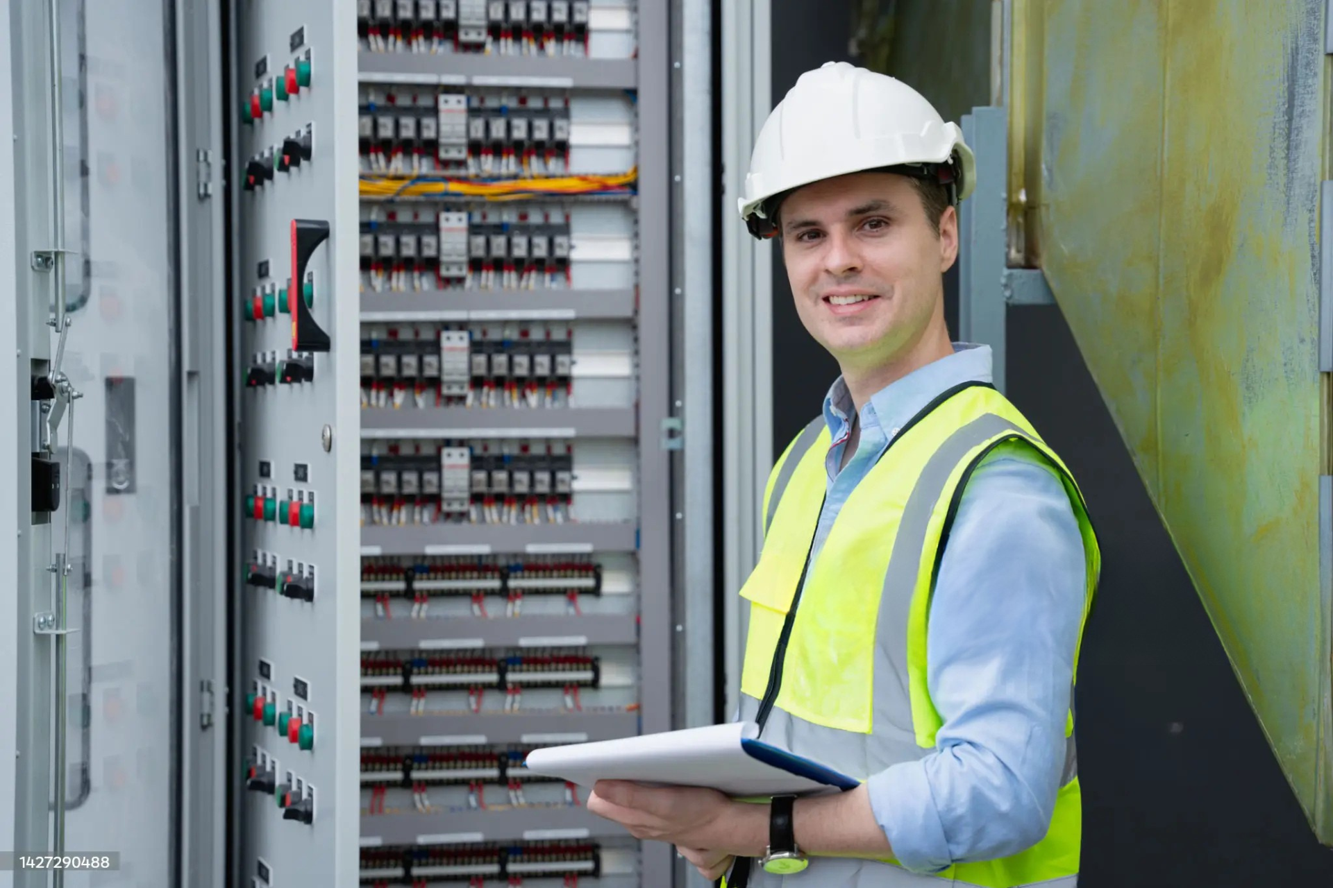 "Electrician wearing a white hard hat and yellow safety vest, holding a clipboard in front of an electrical control panel filled with circuit breakers and colorful wires."