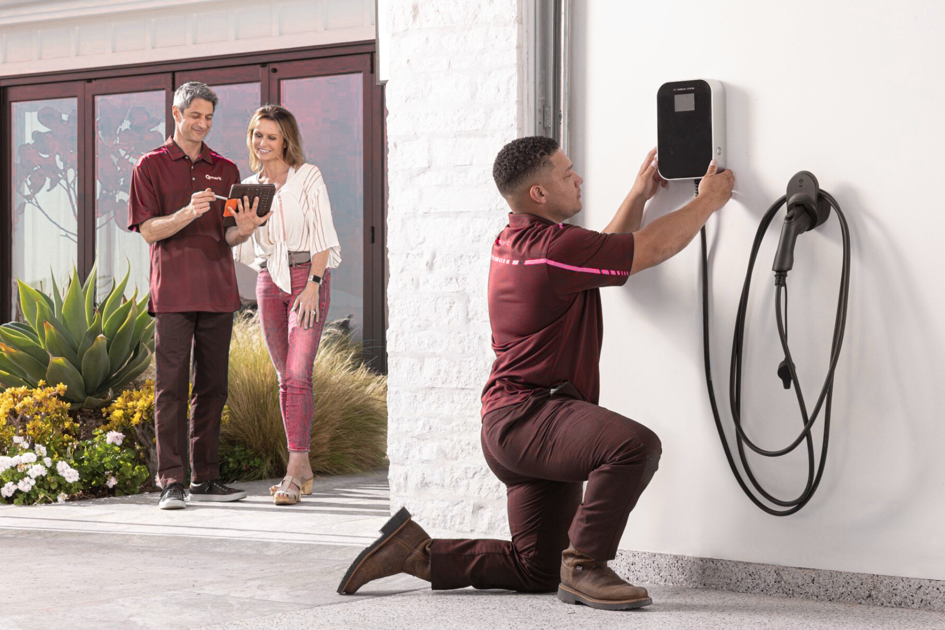 "Electrician installing an EV charging station on a wall while a couple observes and takes notes, showcasing modern home energy solutions."
