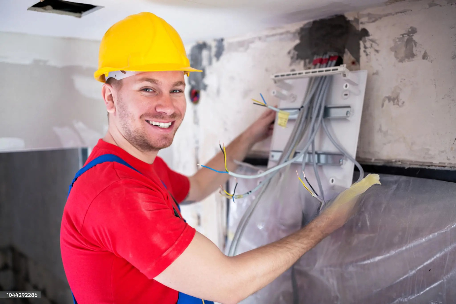 "Electrician smiling and installing an electrical panel with colorful wires, wearing a yellow hard hat and blue overalls in a construction environment."