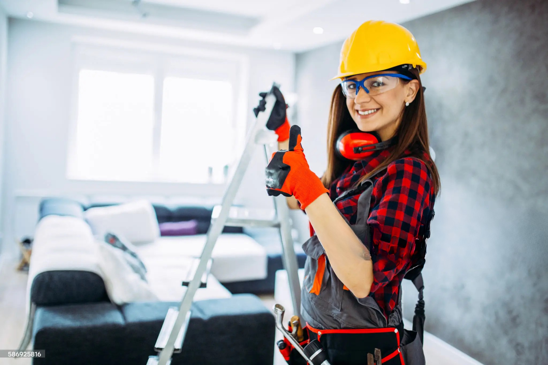 "Female electrician in a yellow hard hat, safety glasses, and gloves, smiling and giving a thumbs-up while standing on a ladder in a modern living room."