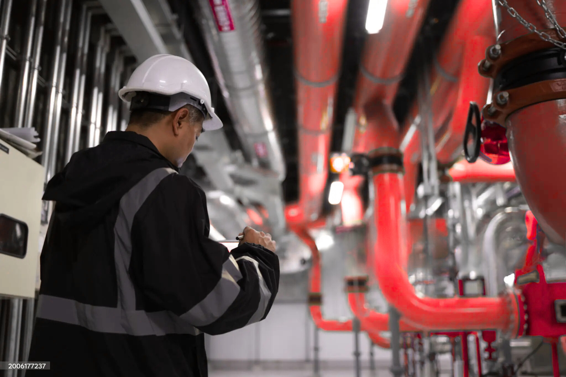 "A worker in a helmet and safety jacket, inspecting pipes in an industrial setting, with bright red pipes running along the ceiling and walls."