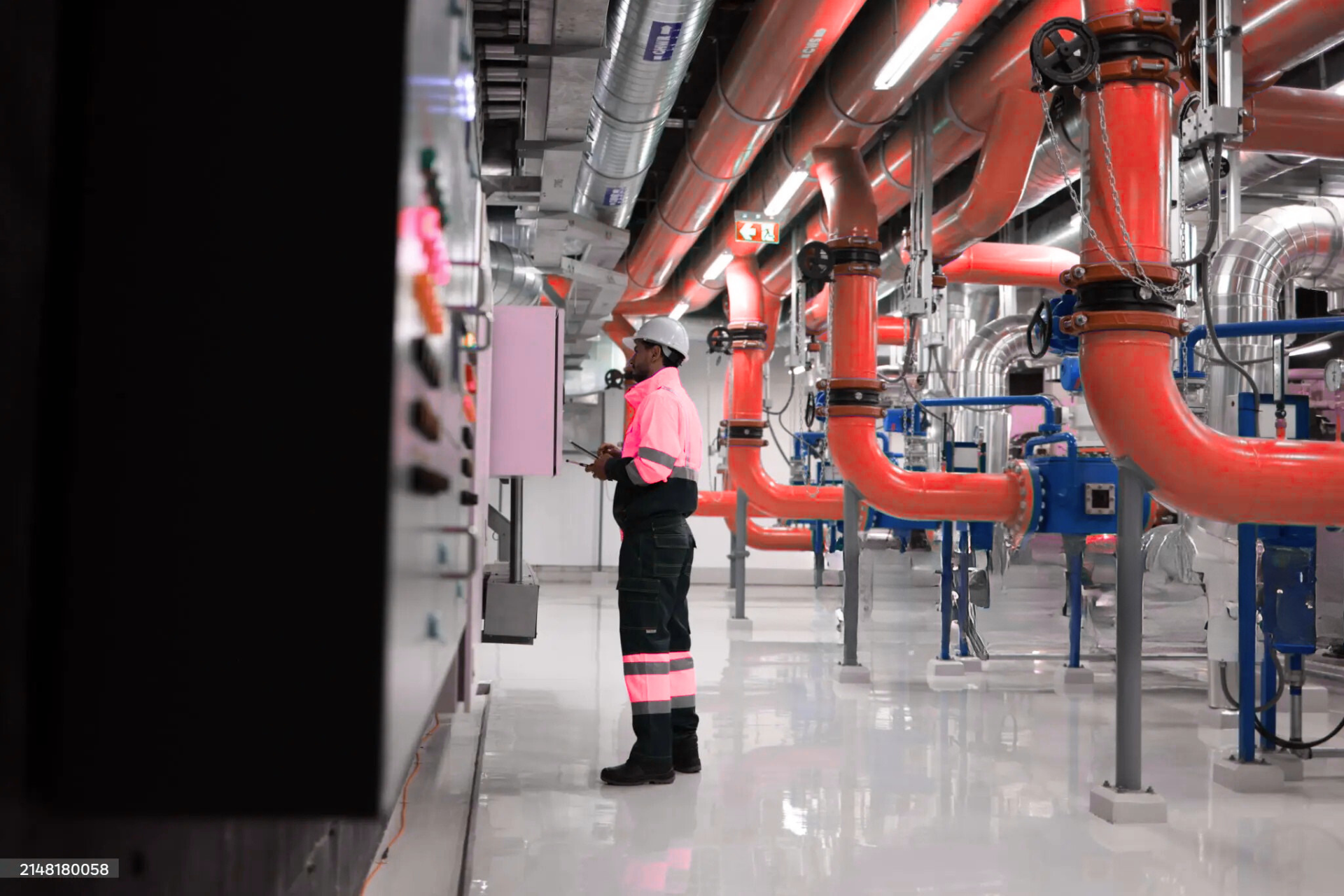 "A worker in a high-visibility pink safety vest and helmet, standing in front of a control panel, with large red pipes and industrial machinery in the background."
