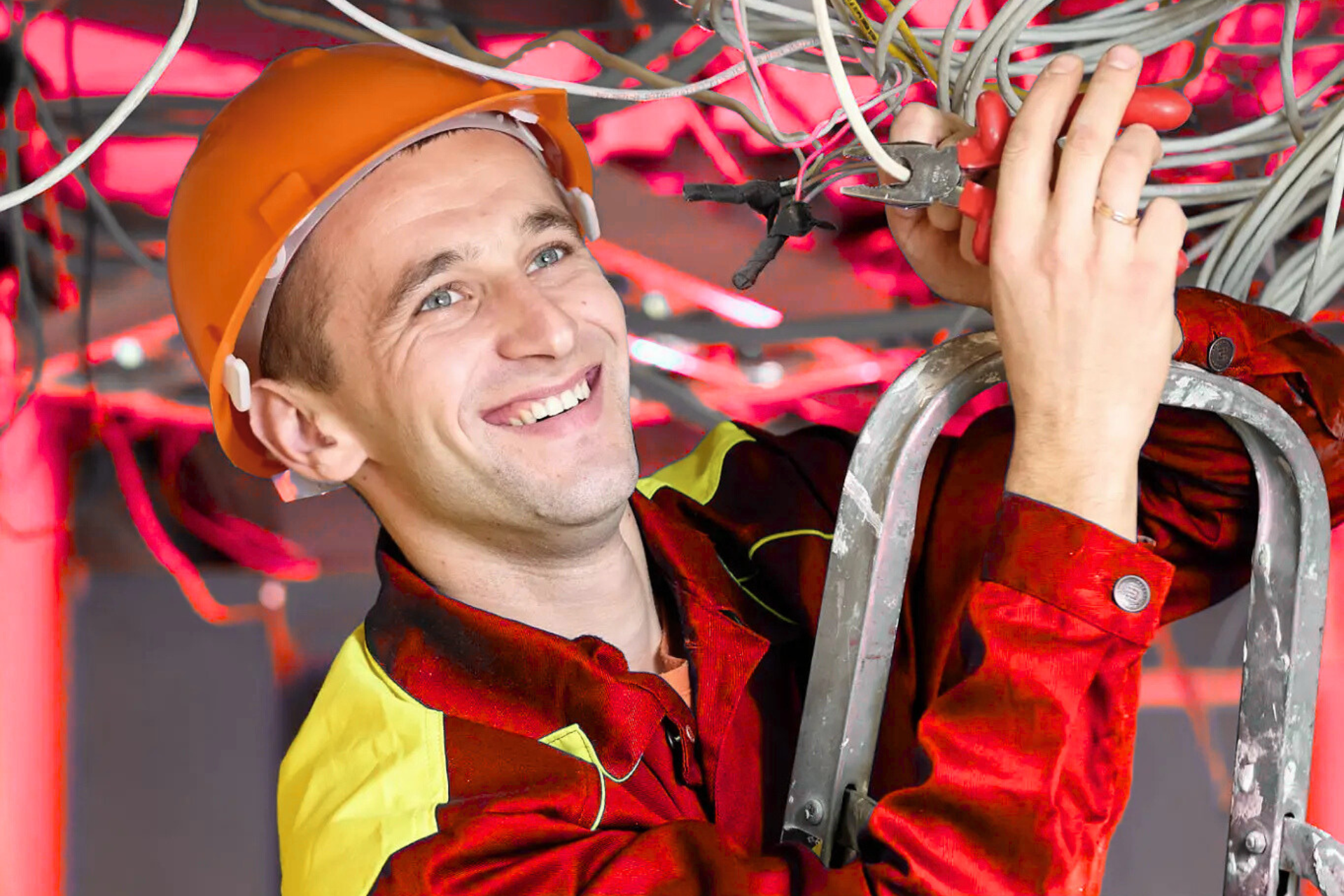 An electrician smiling while working with electrical wires, holding a pair of wire cutters, and wearing an orange helmet and a red work suit, standing on a ladder.