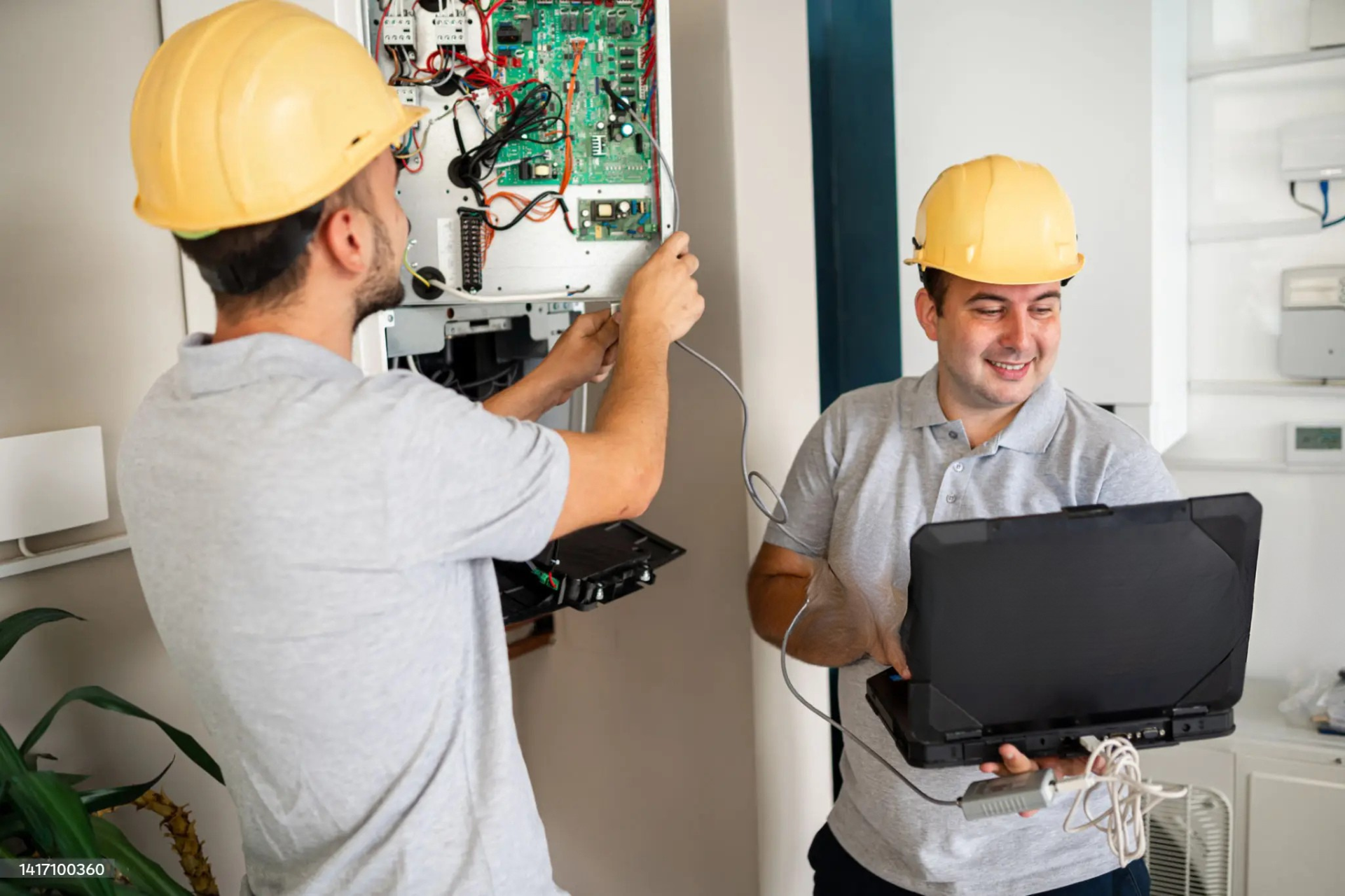 "Two electricians in yellow hard hats working together, one installing wiring into an electrical panel and the other using a laptop to check the system."