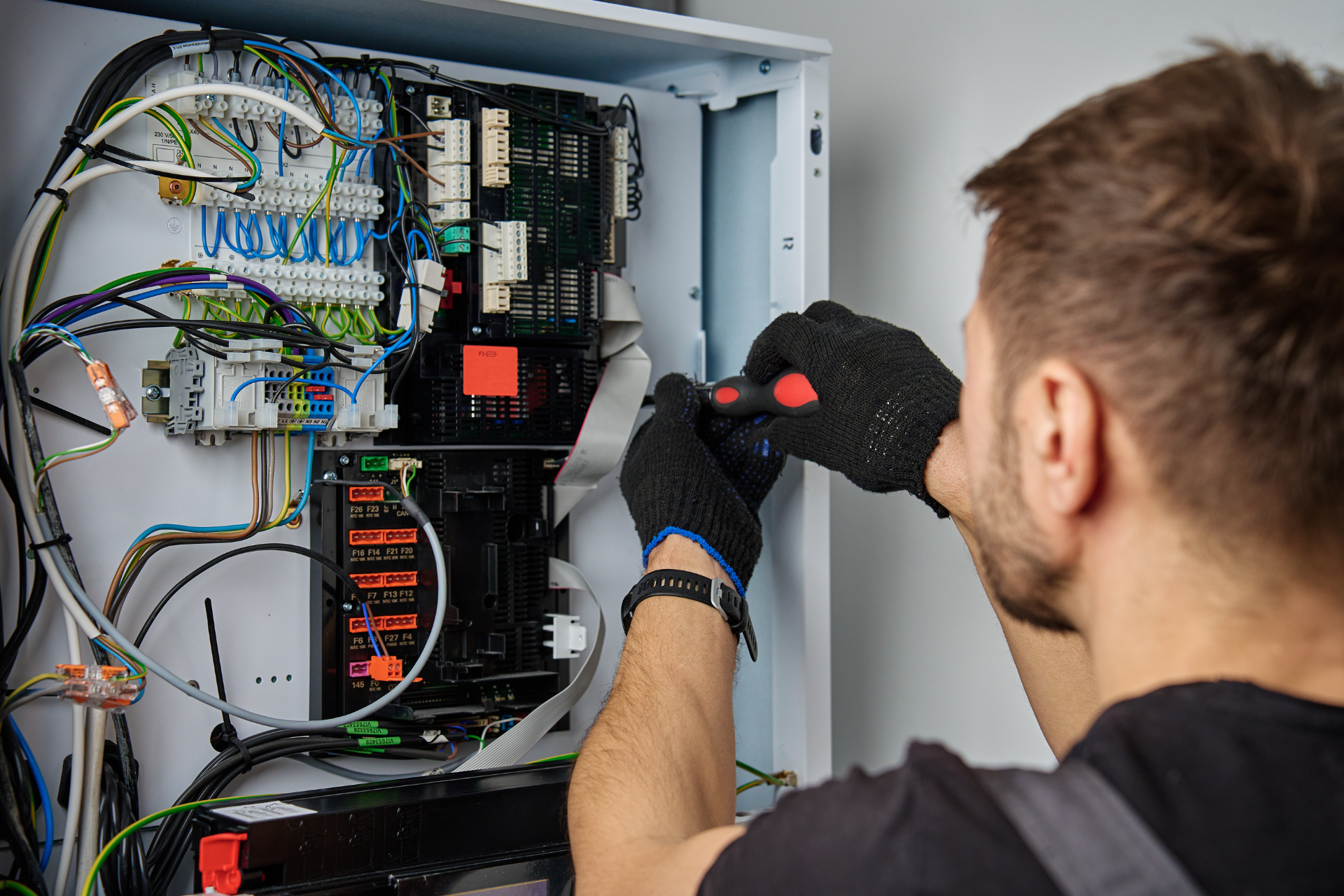 "Electrician wearing black gloves using a screwdriver to work on an electrical panel with colorful wires and terminals."