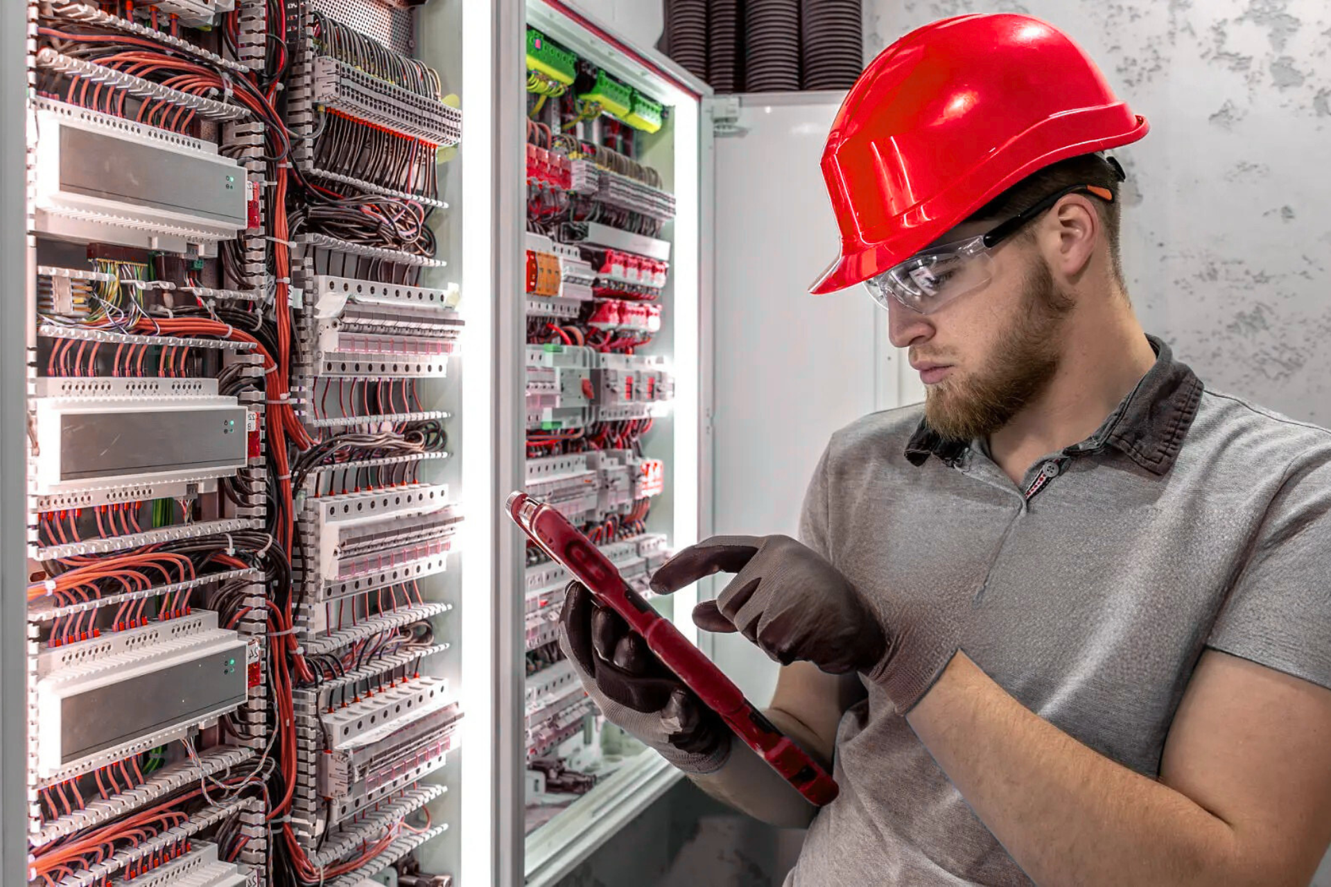 "Electrician wearing a red hard hat and safety glasses, using a tablet to check electrical wiring in a control panel with numerous colored wires."