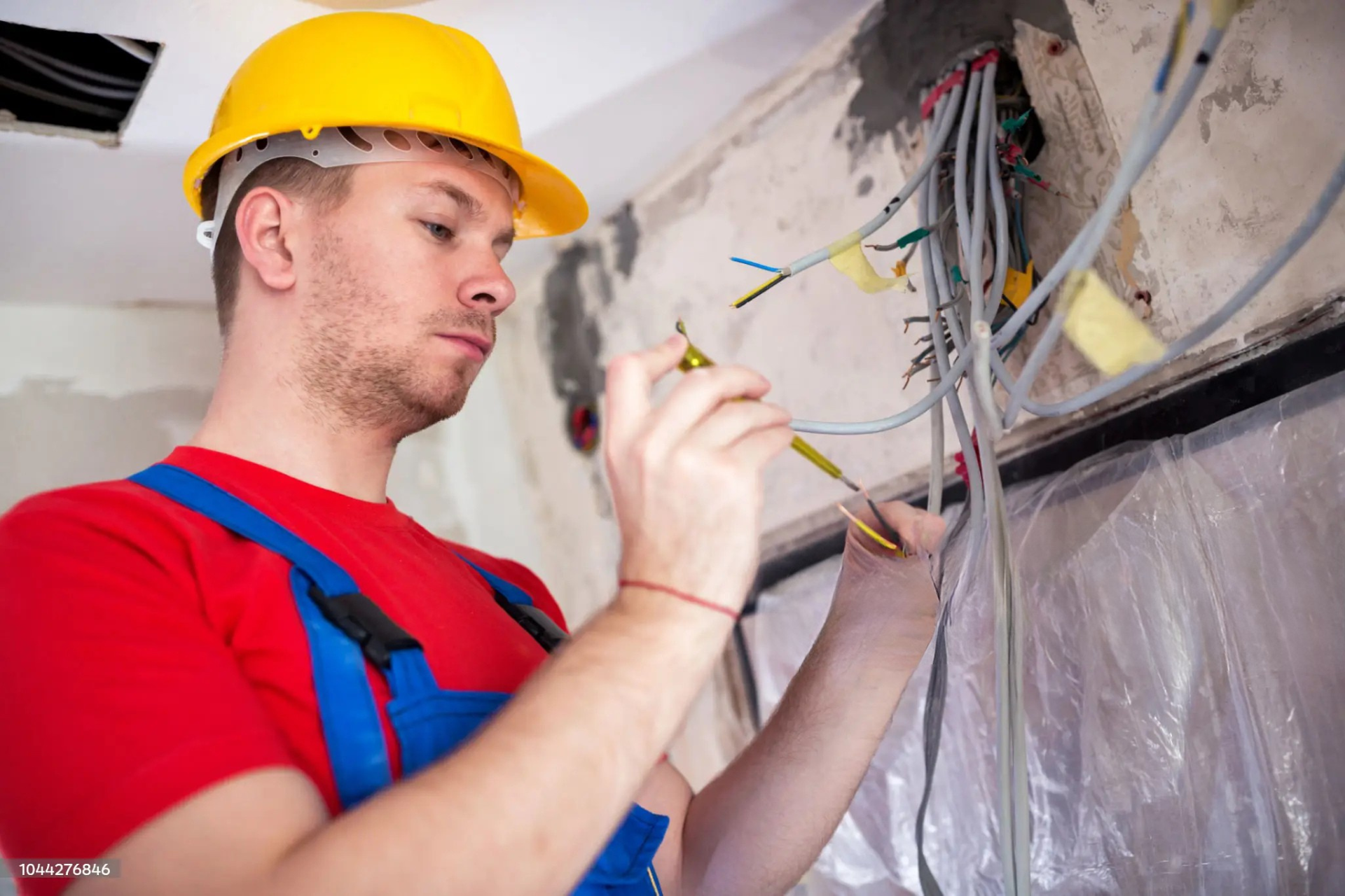 "Electrician in a yellow hard hat and blue overalls working with electrical wires and tools, performing installation or maintenance in a construction site."
