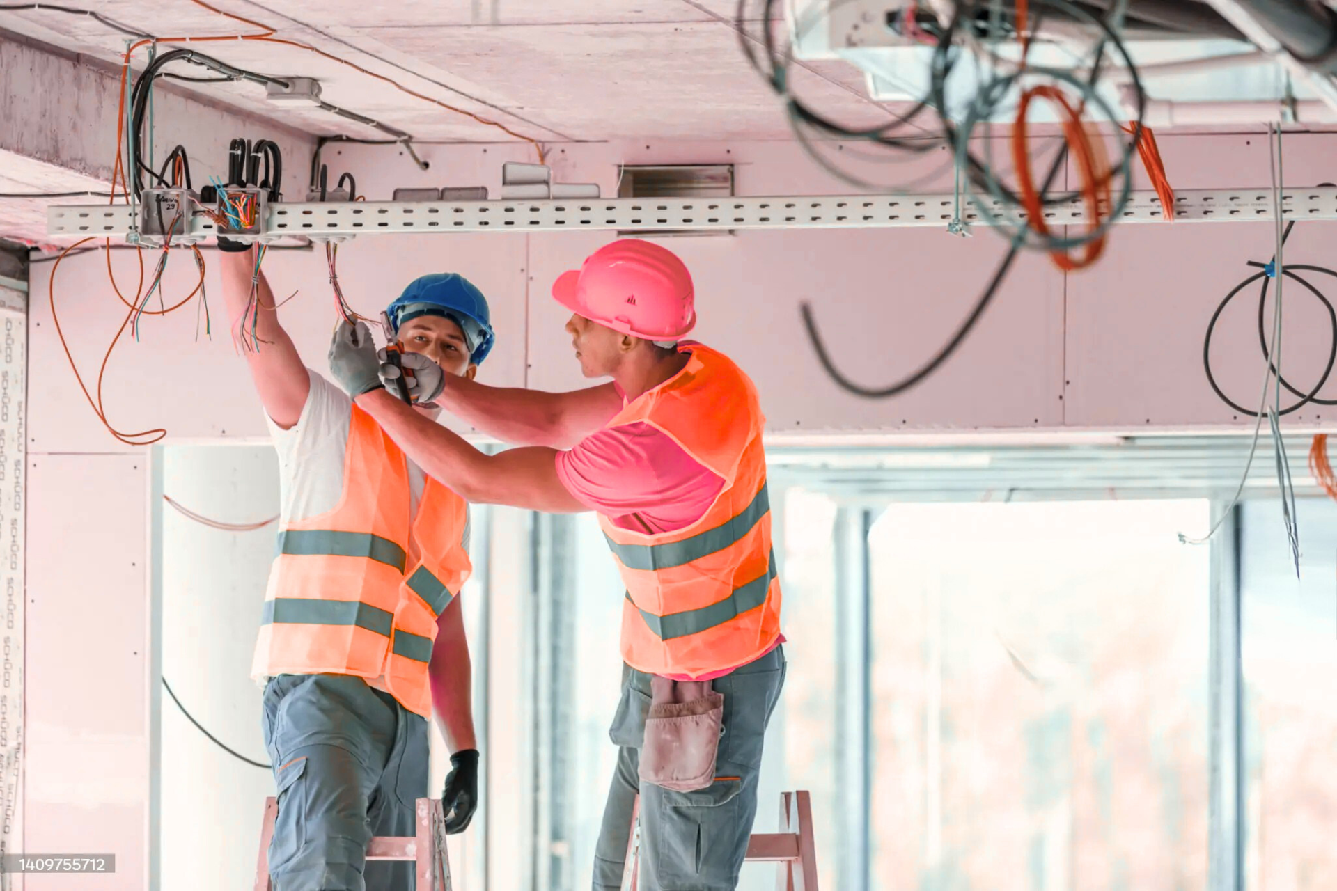 Two construction workers in safety vests and helmets, working together to install electrical wiring on a ceiling in a building under construction.