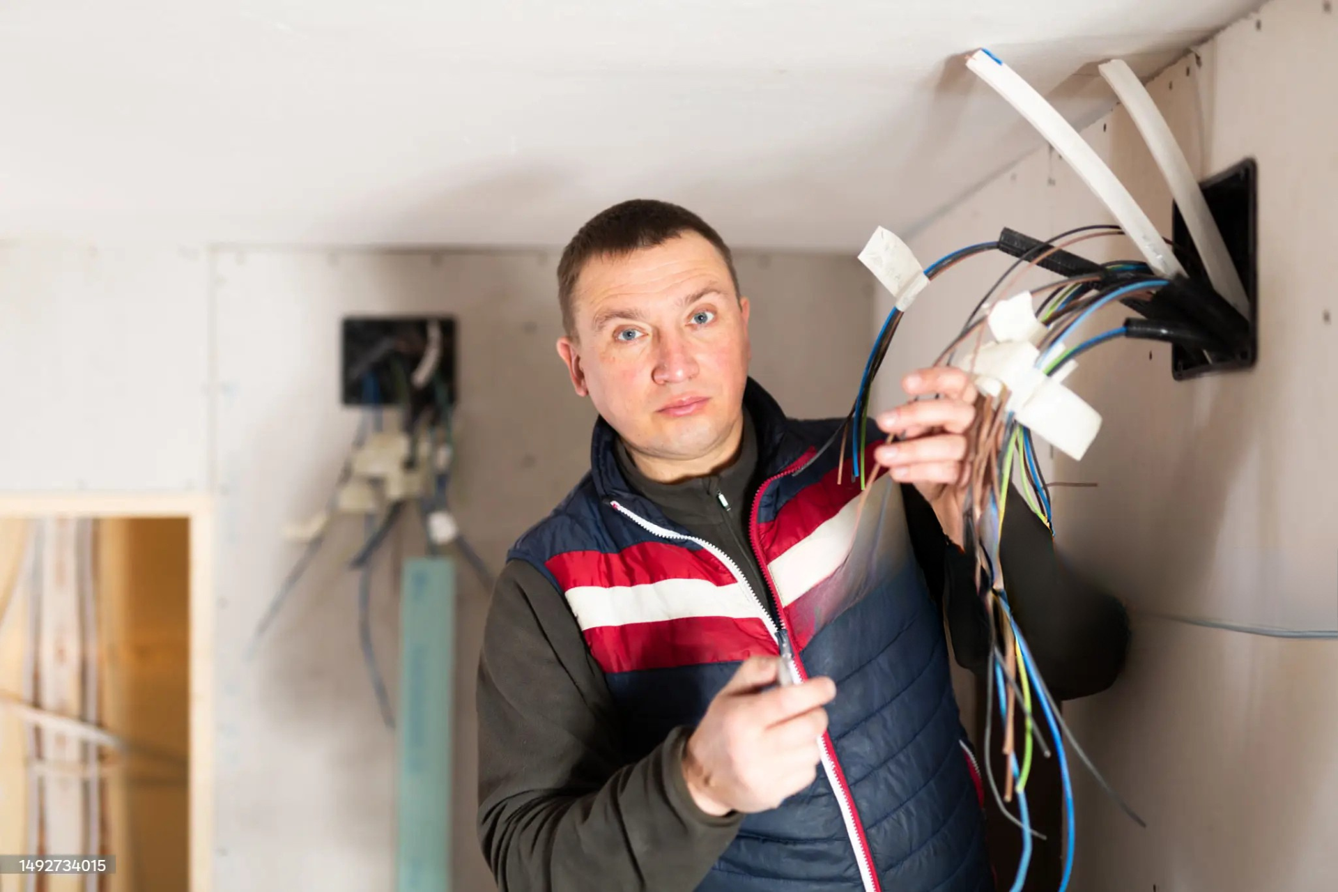 "Electrician holding bundled electrical wires while standing in front of a wall-mounted electrical box, showing the wiring during installation in a construction site."
